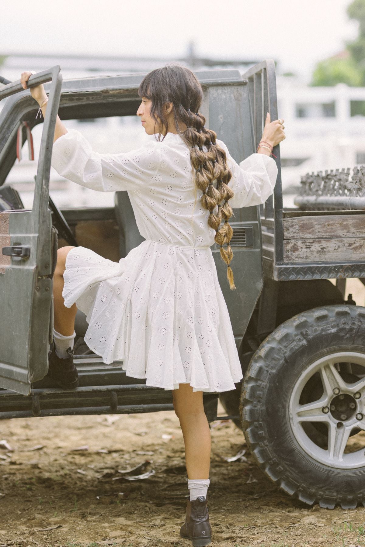 A woman with long braided hair, wearing a La Rosa Dress in vintage embroidered cotton by Myrah Penaloza and a pair of boots, steps into an open jeep. The background reveals a blurred outdoor setting, suggesting adventures that promise carbon-neutral shipping memories.