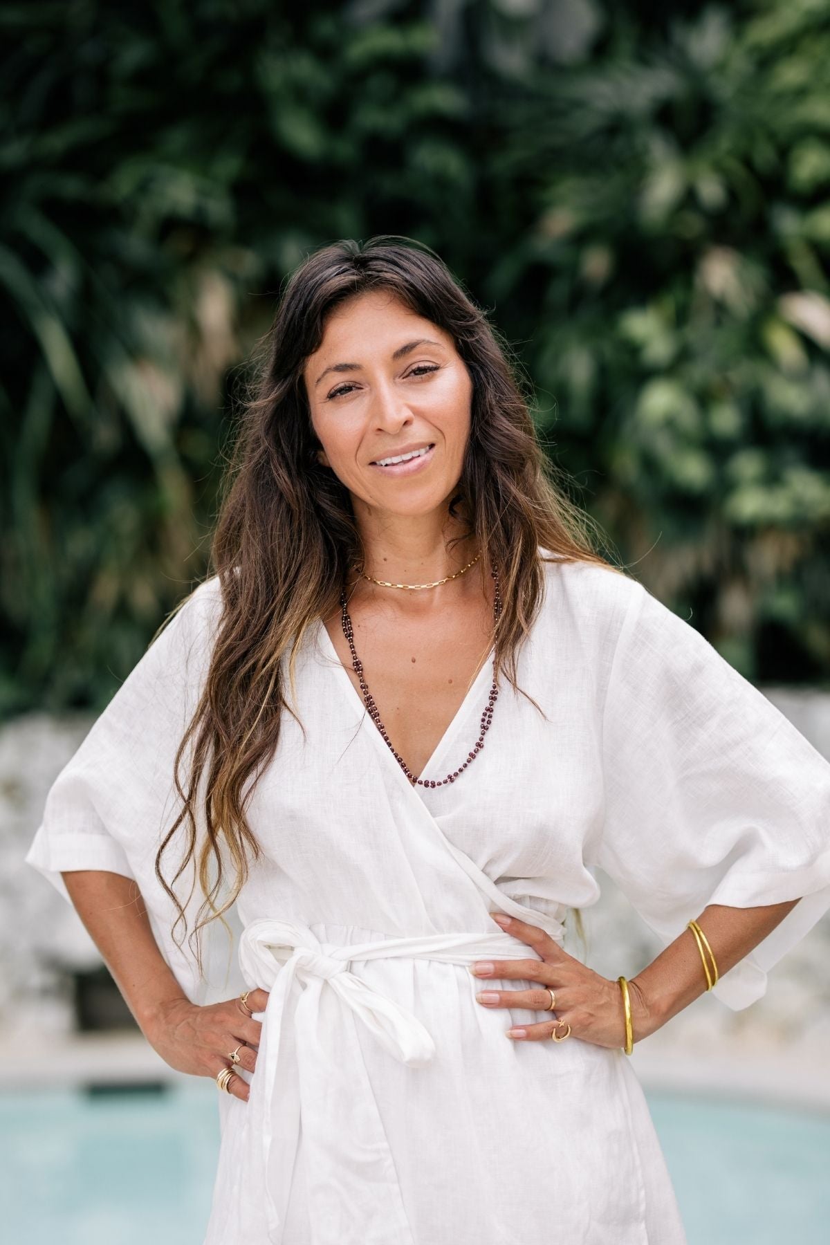 A woman with long brown hair stands outdoors in a 100% linen Magdalena Gown by Myrah Penaloza, adorned with jewelry and smiling gently. The lush greenery in the background enhances the serene and natural setting, highlighting the gown's naturally durable elegance.