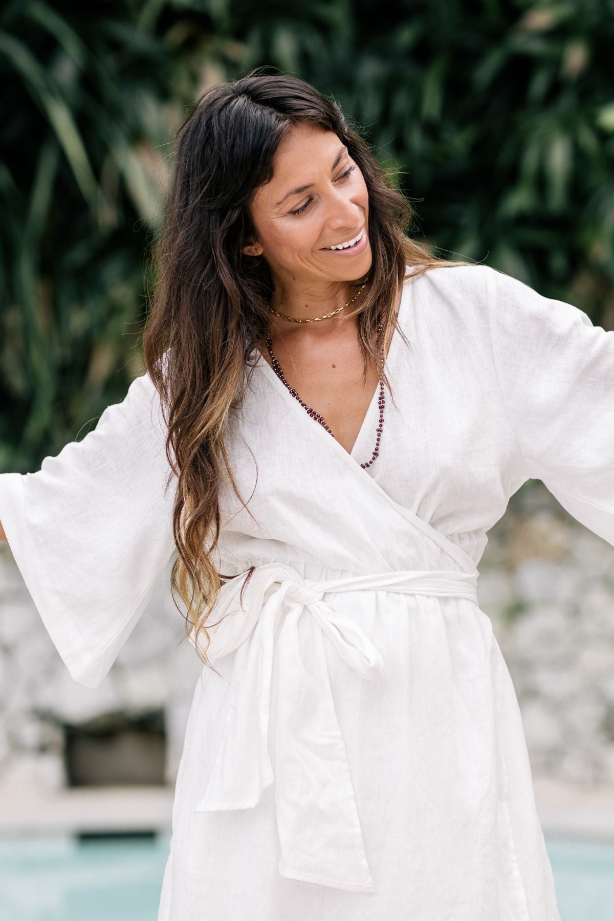 A woman with long brown hair is smiling and gazing to the side, dressed in the Magdalena Gown by Myrah Penaloza, a white moonlight-colored kimono dress made from 100% linen with loose sleeves. She accessorizes with a beaded necklace while surrounded by greenery and blurred elements in an outdoor setting.