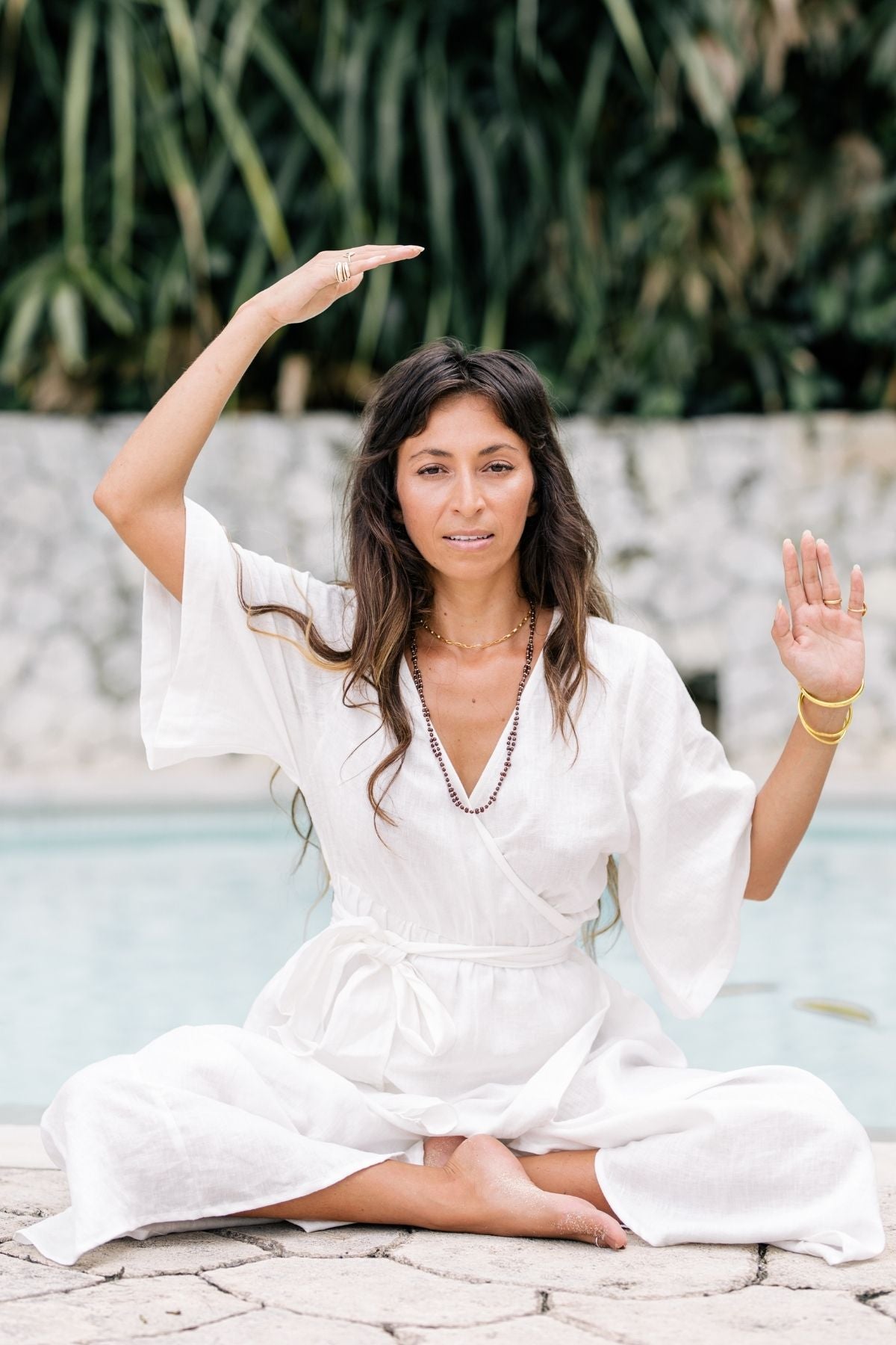 A woman sits cross-legged by a pool, her elegant Magdalena Gown in Moonlight flowing gracefully. Her arms are raised, one hand facing upward and the other sideways. With long brown hair and sparkling jewelry, she is framed by lush greenery in the background. The gown from Myrah Penaloza beautifully complements the serene setting.