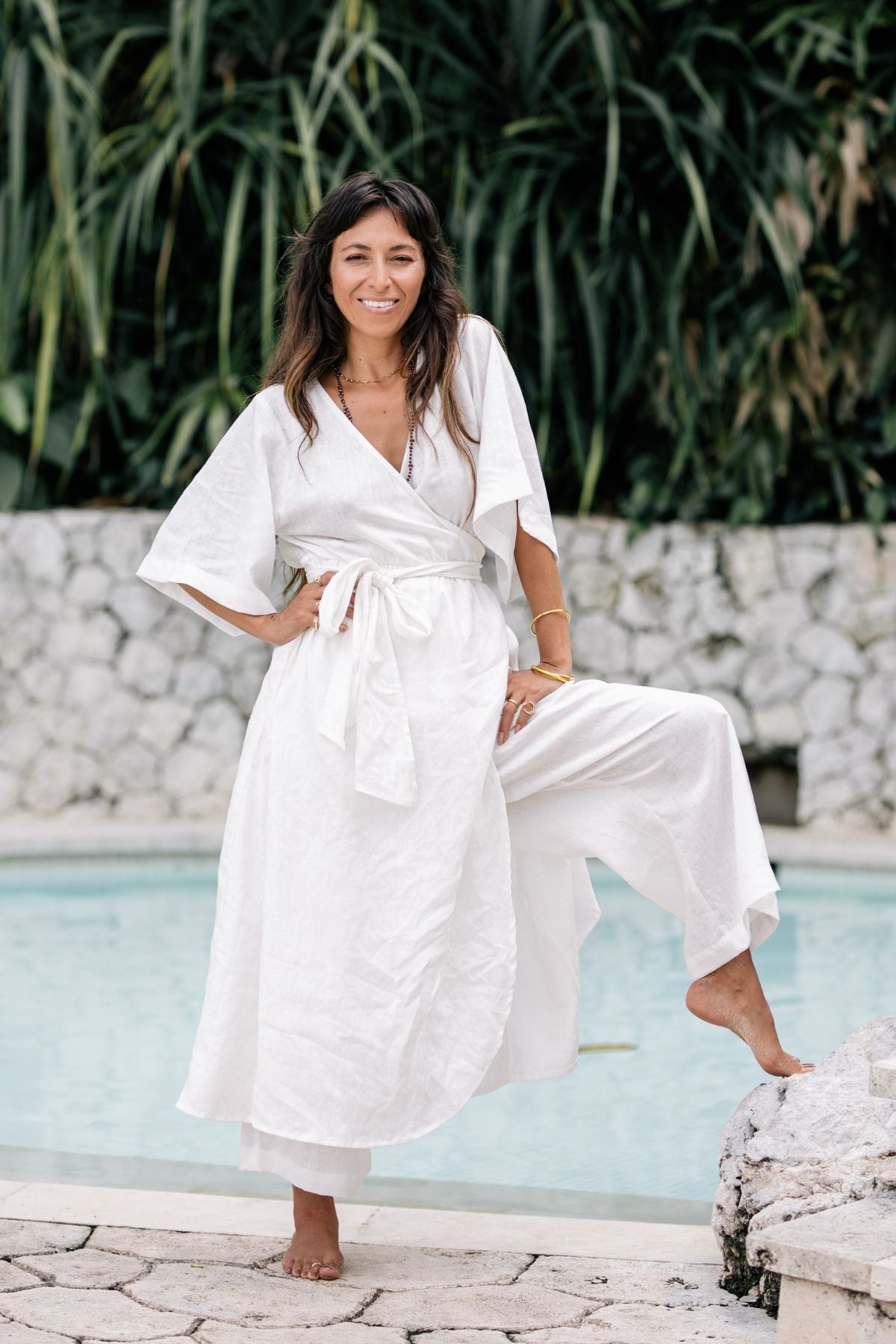 A woman in a Myrah Penaloza Magdalena Gown, crafted from 100% Linen in Moonlight, poses playfully by a pool with one leg resting on a rock. Her long brown hair cascades down as she smiles, surrounded by lush green foliage and a stone wall backdrop.