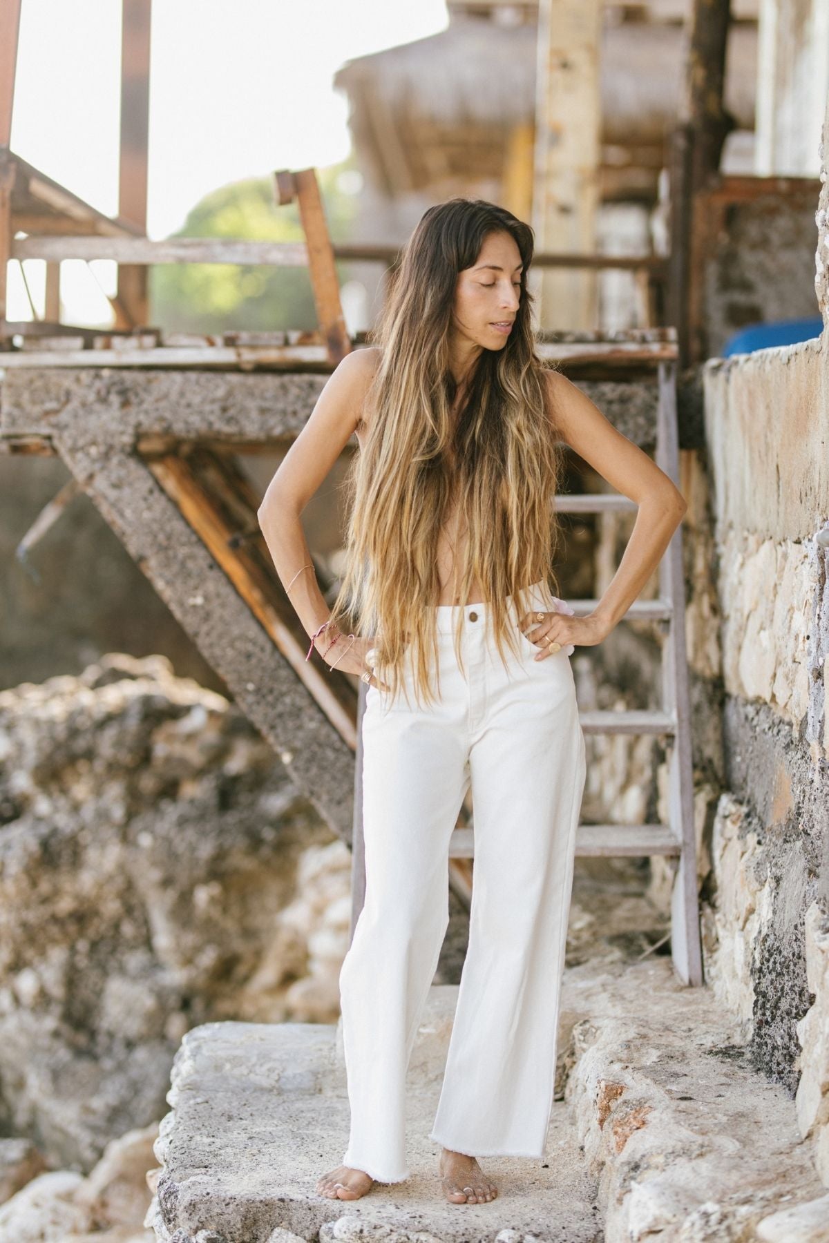 A woman with long, light brown hair stands barefoot on a rocky surface beside a ladder and rustic structure. She wears a sleeveless beige top and the comfortable must-have Miley (High Waisted) Jean, available for pre-order only from Myrah Penaloza. With her hands resting on her hips, she gazes downwards.