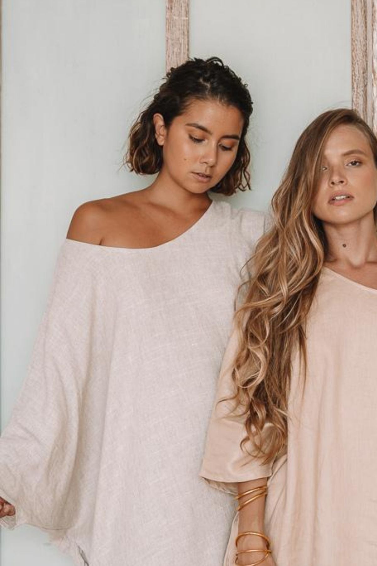 Two women showcase a graceful charm as they stand together against a light backdrop, dressed in Myrah Penaloza's Butterfly Tunic. One has a short, curly hairstyle, while the other wears her hair long and wavy. Their linen attire radiates ease and sophistication, complemented by subtle jewelry choices.