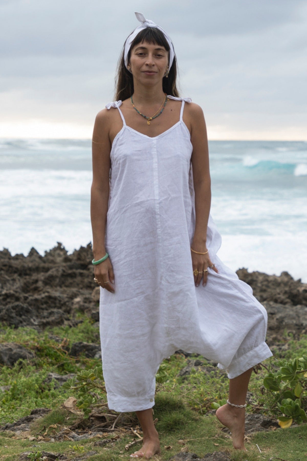 Standing barefoot on rocky terrain by the ocean, a person wears the loose-fitting, 100% linen Japa Playsuit from Myrah Penaloza along with a headband. The cloudy sky and ocean waves in the background create a relaxed, beach-side appearance evocative of Kundalini Yoga serenity.