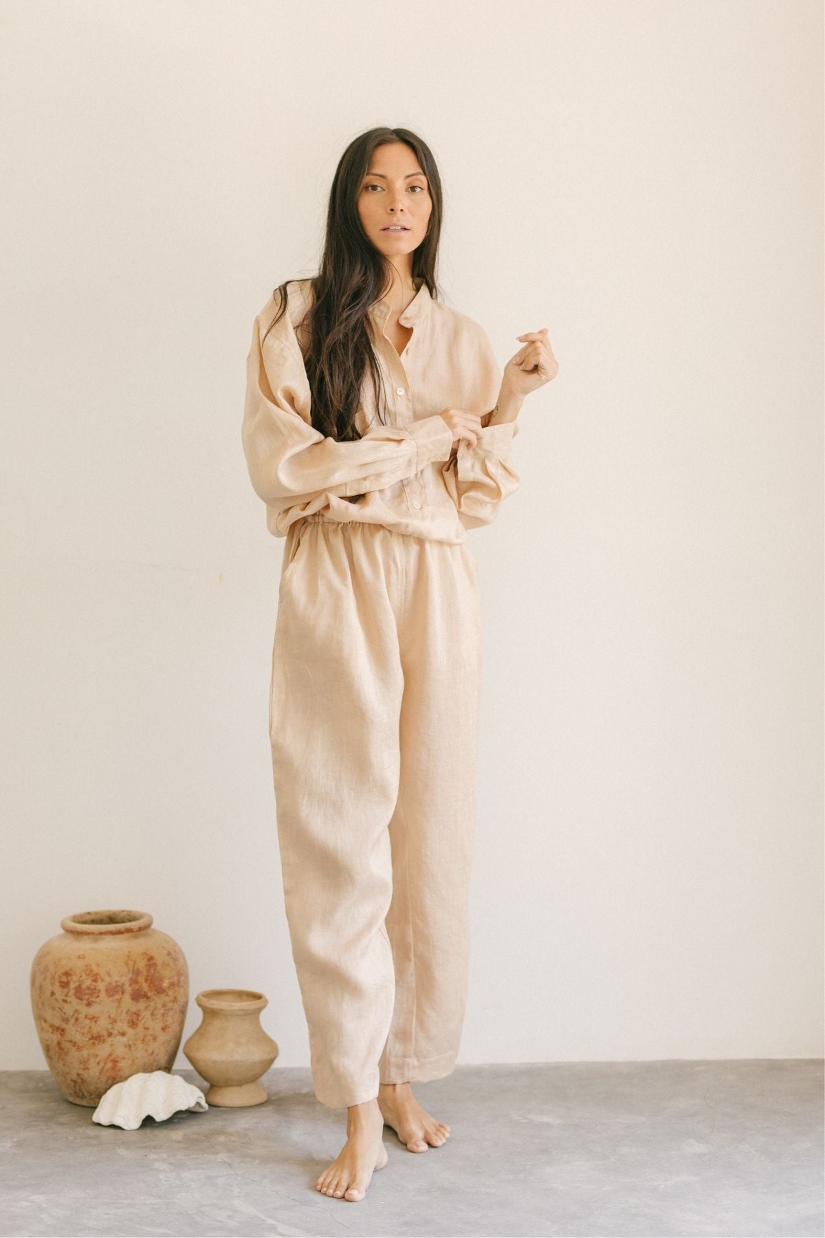 A woman with long dark hair stands barefoot on a concrete floor, wearing the Gather Playsuit in Light Linen and Clay by Myrah Penaloza. In front of the neutral wall, two ceramic vases and a shell accompany the serene scene, echoing the simplicity of stone-washed linen style.