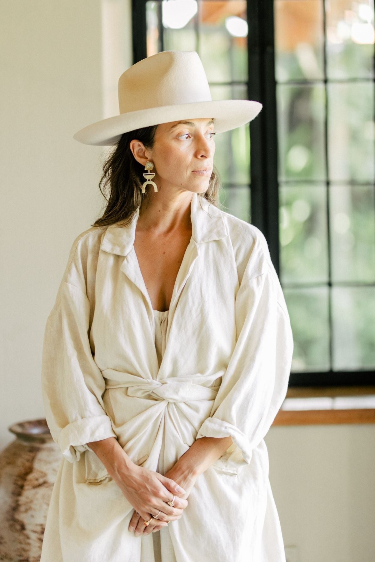 Wearing an Infinity Jacket by Myrah Penaloza, made from 100% linen, a person stands by a window in thought, adorned with a wide-brimmed hat and large earrings. Their light and loose-fitting attire features a knotted front. The softly lit background reveals greenery outside—a perfect moment from the Moonlight Collection.