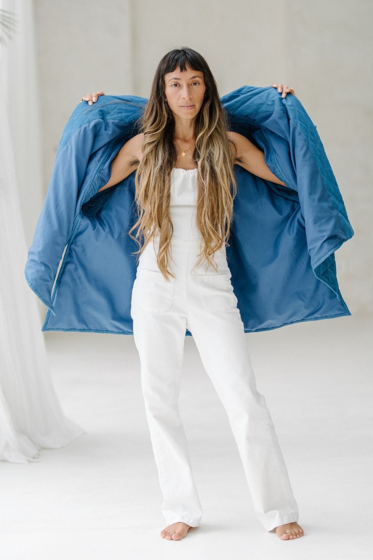 A woman with long hair stands barefoot on a white floor, wearing a white jumpsuit. She holds open an oversized Javi Quilted Jacket (Pre-Order) by Myrah Penaloza, showcasing its inner lining. The background is minimalistic with soft, neutral tones.