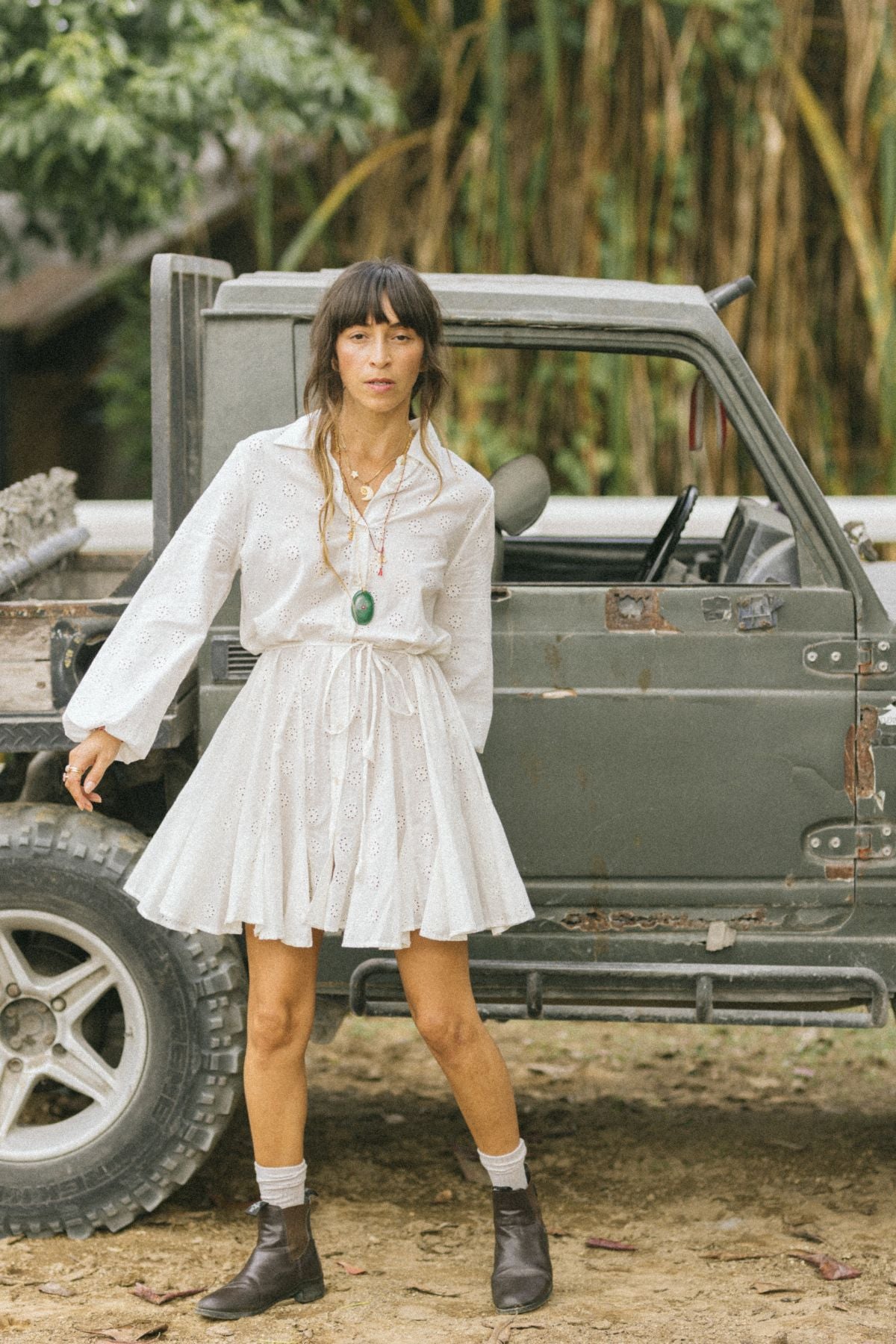 A person wearing a Myrah Penaloza La Rosa Dress, crafted from vintage embroidered cotton, stands beside an old green vehicle outdoors. Sporting long dark hair, the individual enhances their look with brown boots and a green pendant necklace. The setting is enriched by trees and foliage, capturing the essence of natural charm infused with vintage cotton elegance.