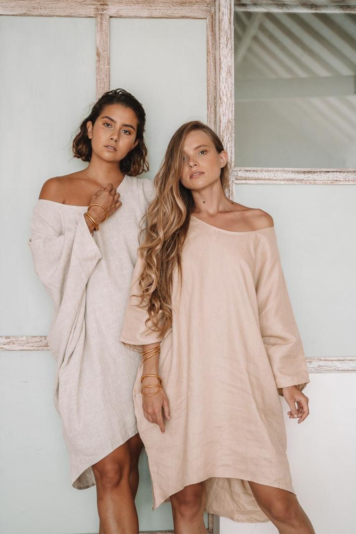 Two women stand in front of a wooden door, embodying the grace of linen clothing. They wear the loose, off-shoulder Butterfly Tunic by Myrah Penaloza in beige tones, perfect as an everyday piece. One has short dark hair, and the other has long light hair; gold bangles adorn their wrists.