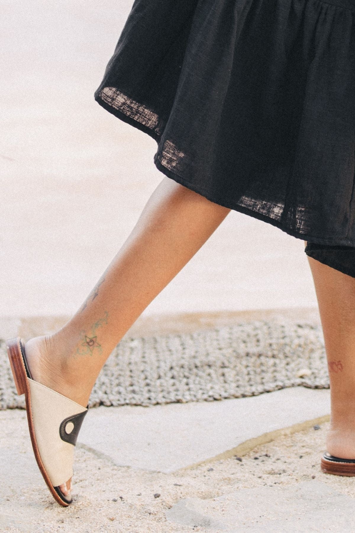 A person wearing a black skirt and the Yin Yang Open Toe Flats from Myrah Penaloza, crafted in a family-run workshop, strolls outdoors, displaying small colorful tattoos on their ankle. The ground is a combination of stone and woven mat.