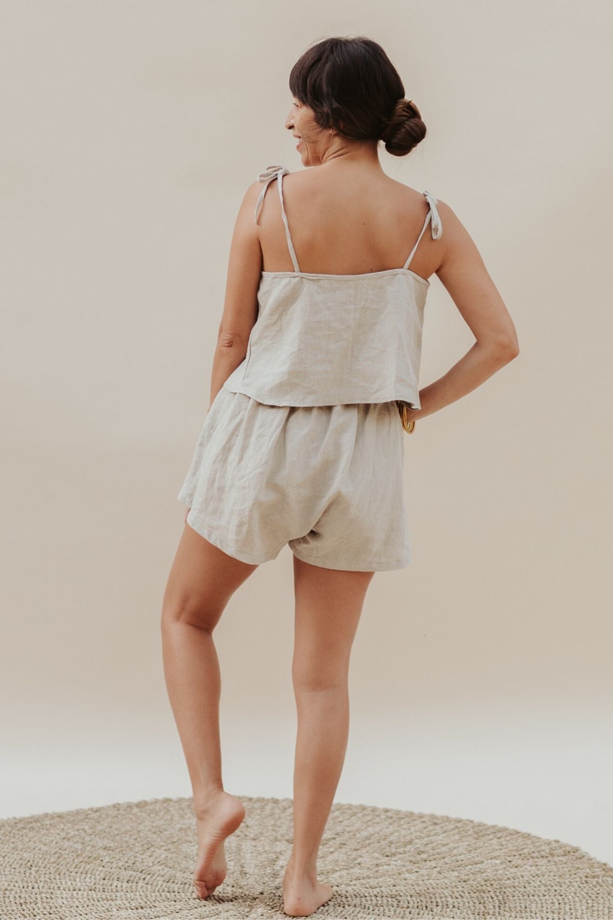 A woman stands barefoot on a woven rug, facing away, wearing the Myrah Penaloza Nidra Sleeper Top Sleeper in clay. The light beige ensemble complements the calming, neutral tone of the background.