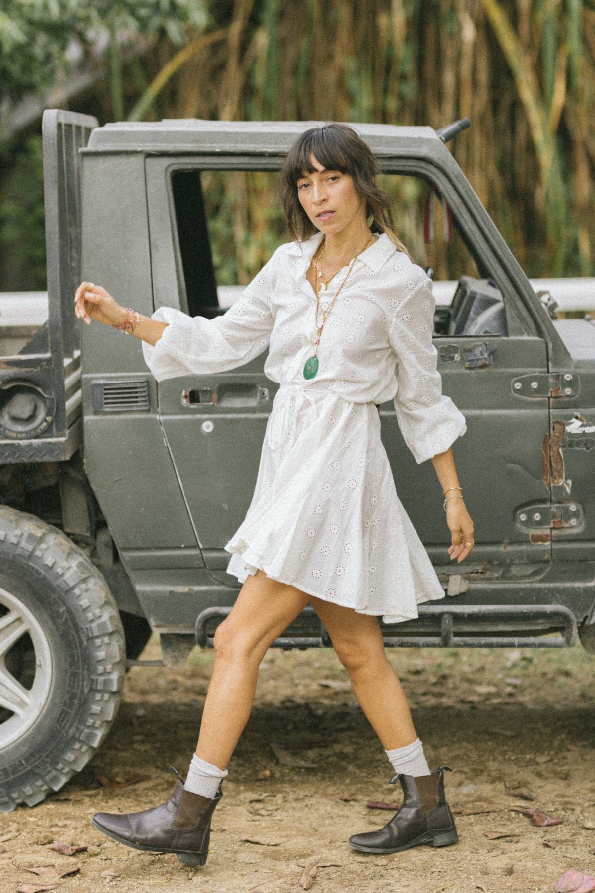 A woman in a vintage La Rosa Dress by Myrah Penaloza stands next to a rugged off-road vehicle, wearing brown boots. Surrounded by lush green foliage, she is adorned with a necklace and bracelets, and her right foot is slightly raised, embodying the spirit of adventure in sustainable style.