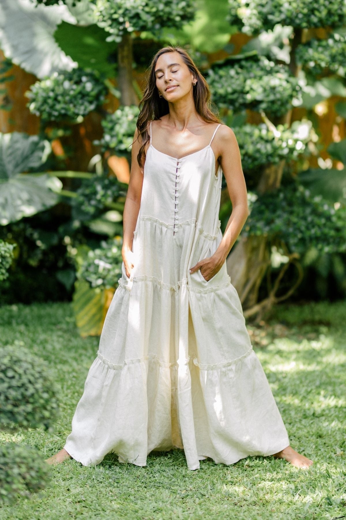 A woman stands outdoors on grass, wearing a long, flowing white creation by Myrah Penaloza, crafted from sustainable 100% linen. She has long brown hair and her eyes are closed, exuding a sense of relaxation. Lush green plants and trees surround her against the serene backdrop of nature's embrace under the moonlight.