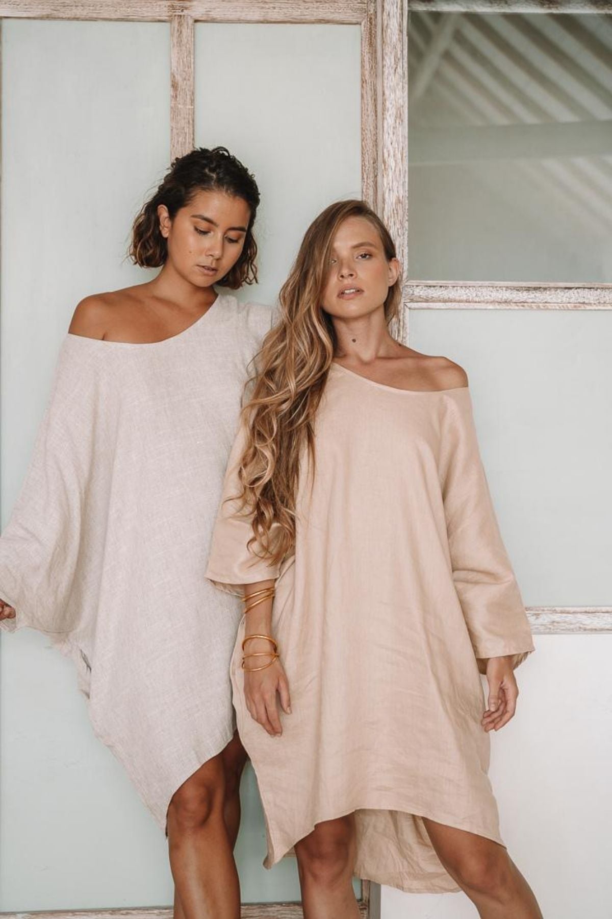 Two women stand side by side in effortless Butterfly Tunics from Myrah Penaloza, wearing off-shoulder, loose-fitting dresses in neutral tones. They pose casually in front of a rustic wooden door with frosted glass panels, their relaxed expressions and long, wavy hair adding charm to this everyday piece.