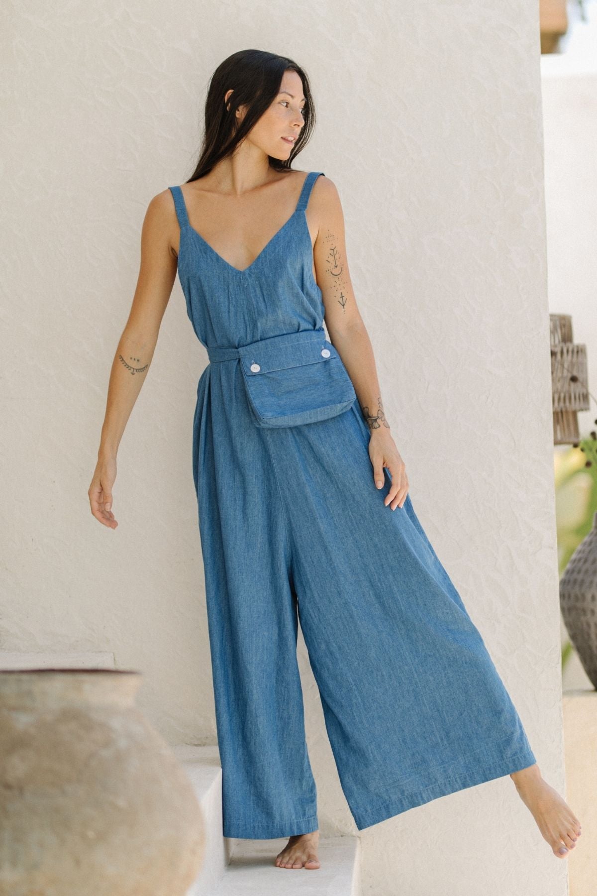 A woman with long dark hair stands barefoot on a step, wearing a Denim Stonewashed Kundalini Playsuit by Myrah Penaloza, complete with a pocket belt. She has tattoos on her arms and gazes to the side against a background of a light-colored wall and decorative pots.