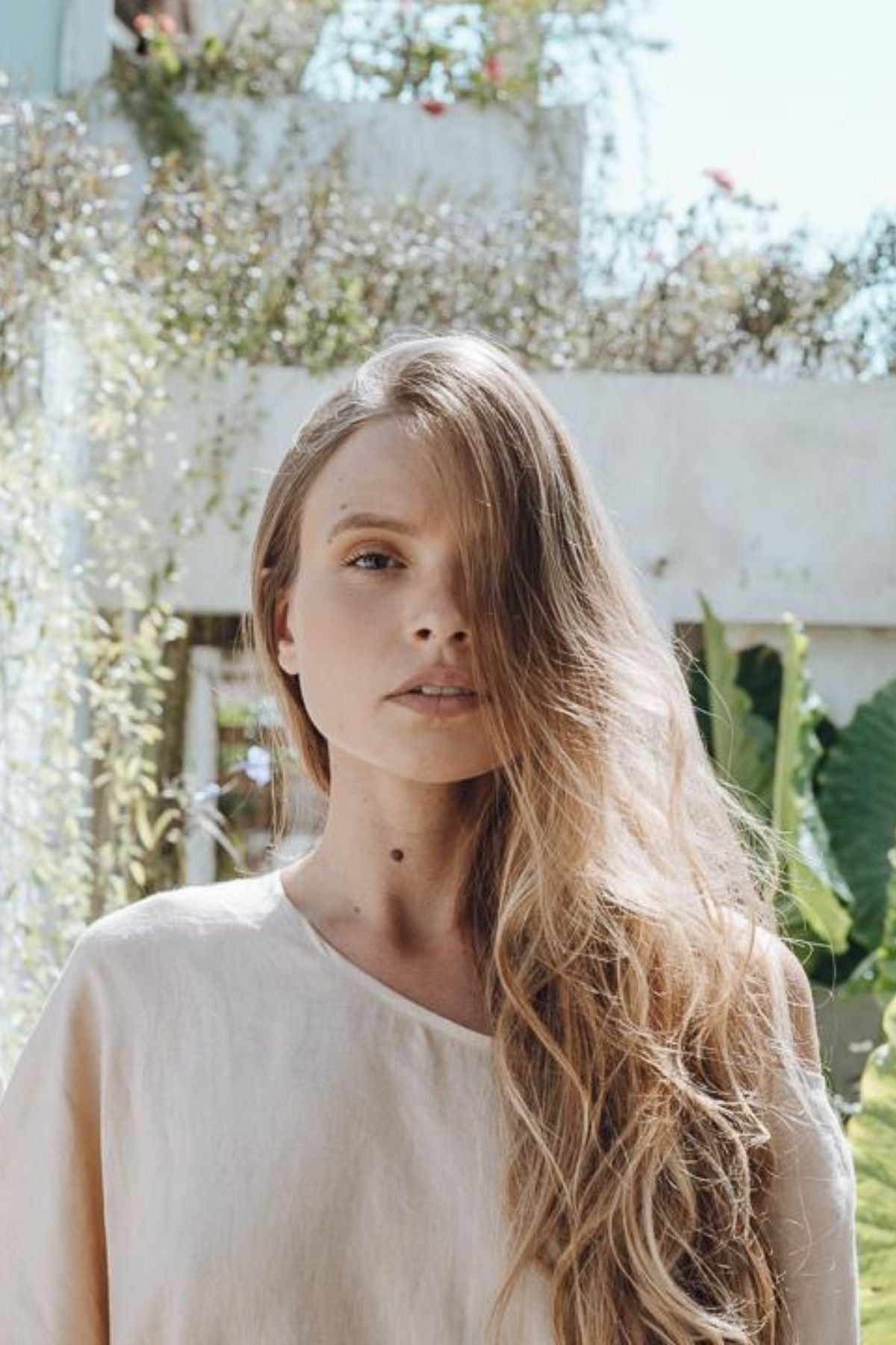 In a sun-drenched garden, a woman with cascading hair exudes elegance in the Butterfly Tunic by Myrah Penaloza. She is surrounded by lush greenery and a white structure, with gentle sunlight enhancing the tranquil atmosphere.