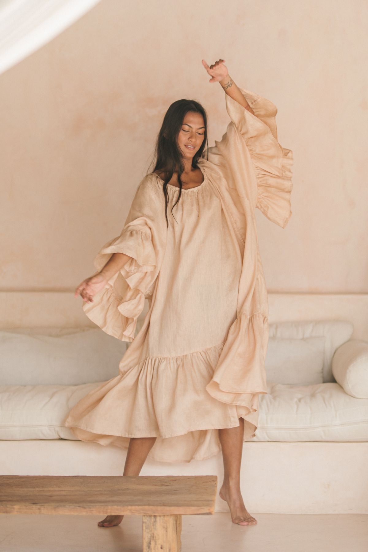 A woman with long hair joyfully dances in a Myrah Penaloza Brida Linen Gown Long, featuring ruffled sleeves and hem. She is in a minimalist room with neutral tones, including a white couch and wooden table, reflecting the gown's light beige beauty.