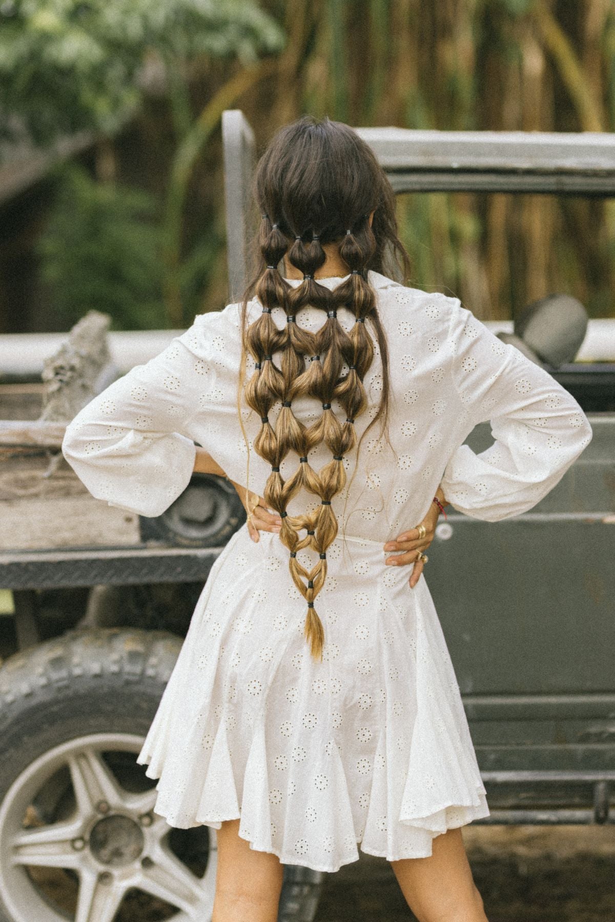 A woman with long, intricately braided hair stands with her back to the camera, wearing the La Rosa Dress from Myrah Penaloza, a vintage embroidered cotton piece. She is in an outdoor setting near a vehicle, surrounded by trees in the background.