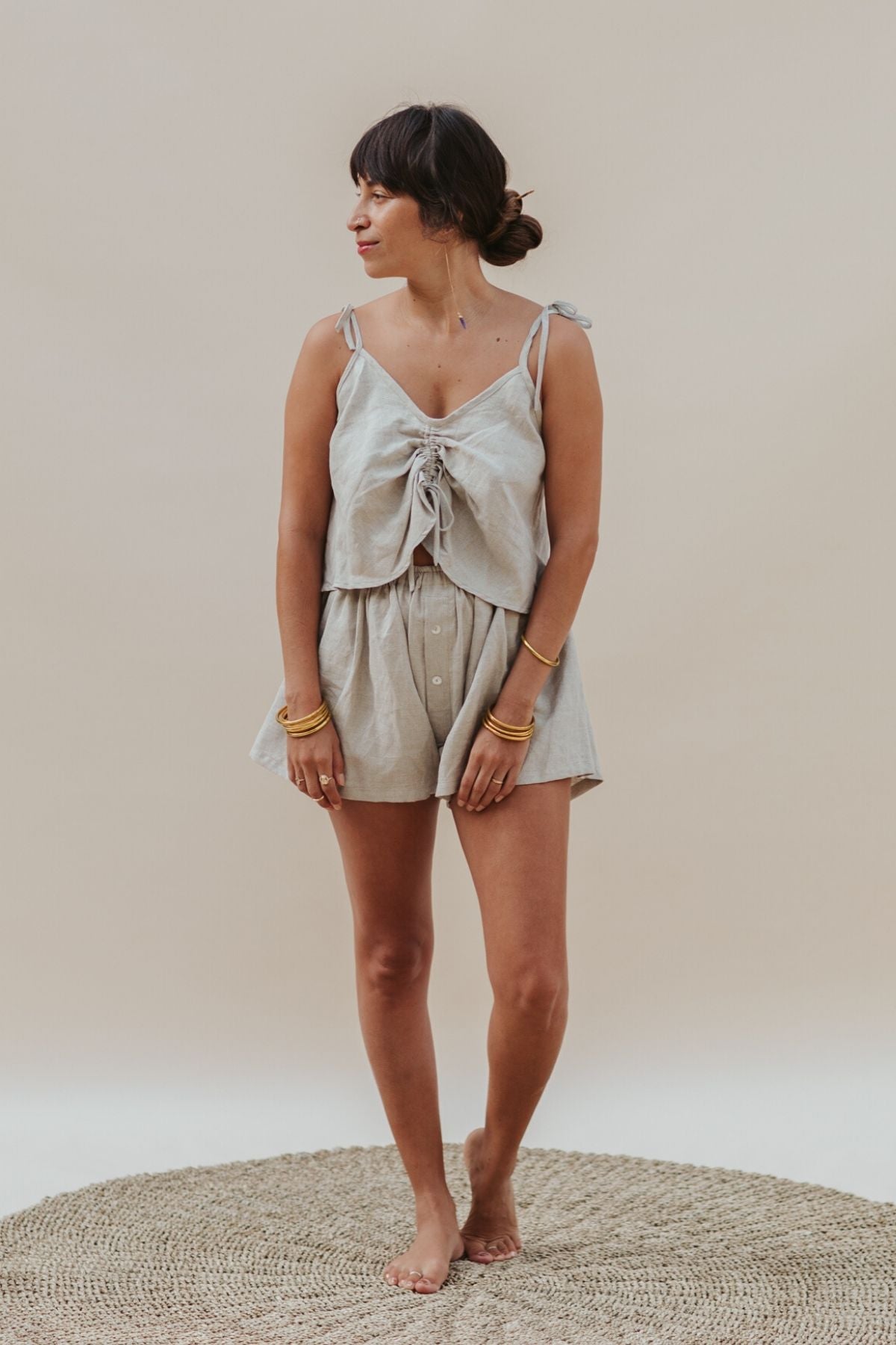 A woman stands barefoot on a round woven rug, wearing the Myrah Penaloza Nidra Sleeper Top in Clay and matching shorts. Her long dark hair is tied back, and she turns her head to the side, with several gold bangles adorning her wrists.