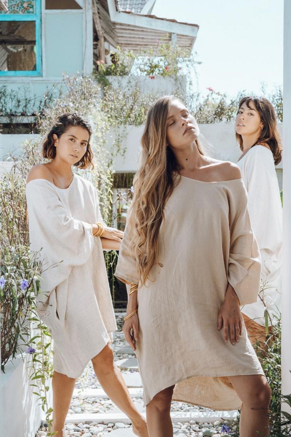 Three women stand outdoors in a garden setting, each wearing the Butterfly Tunic by Myrah Penaloza. They pose confidently among greenery and flowers, with their everyday pieces gracefully illuminated by the sunlight casting a soft glow around them.