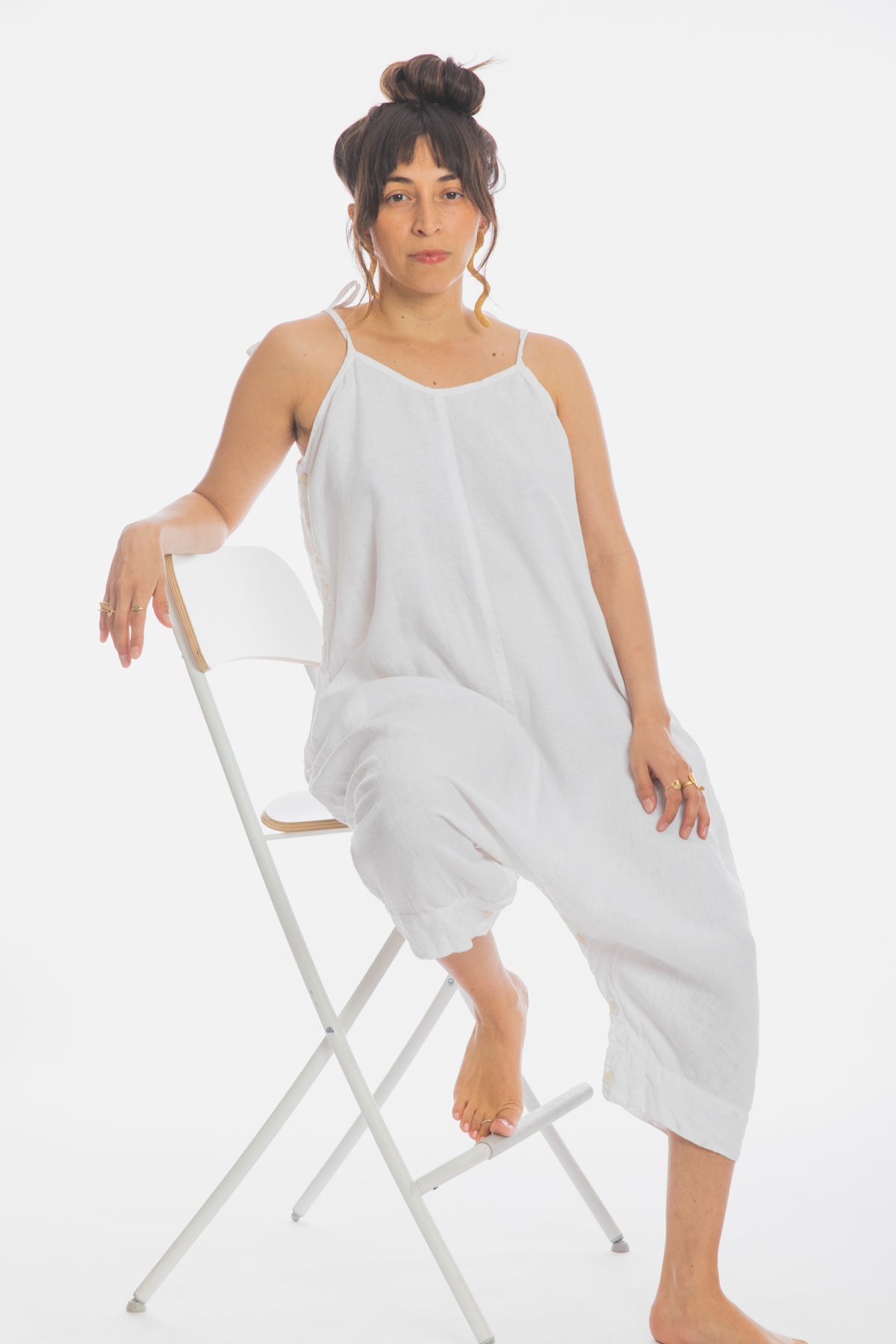 A woman with dark hair in a bun is sitting on a white folding chair. She is wearing a white, loose, sleeveless Japa Playsuit (100% Linen) WI by Myrah Penaloza and has a relaxed expression. One foot rests on the chair while the other touches the ground. The plain white background evokes the serenity of Kundalini Yoga.