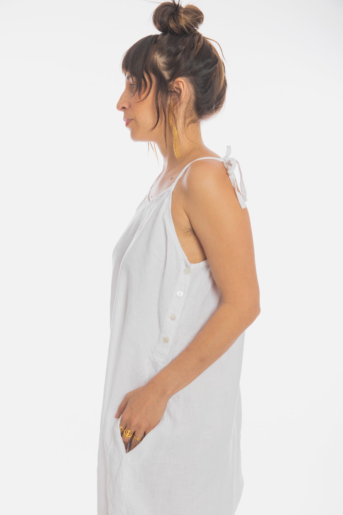 A person with long brown hair tied in a bun is wearing the Japa Playsuit by Myrah Penaloza, a loose-fitting and sleeveless dress made of 100% linen in white, featuring buttons down the sides. They are standing in profile against a white background, holding a small grey object in their hand and embodying serenity akin to those practicing Kundalini Yoga.