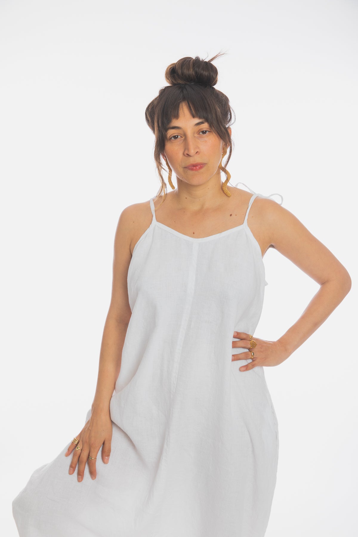 A person with dark hair tied in a high bun is wearing the Japa Playsuit, a loose, white sleeveless outfit made from 100% linen fabric by Myrah Penaloza. They have a serious expression and are standing against a plain white background, with one hand on their hip and the other holding the playsuit.