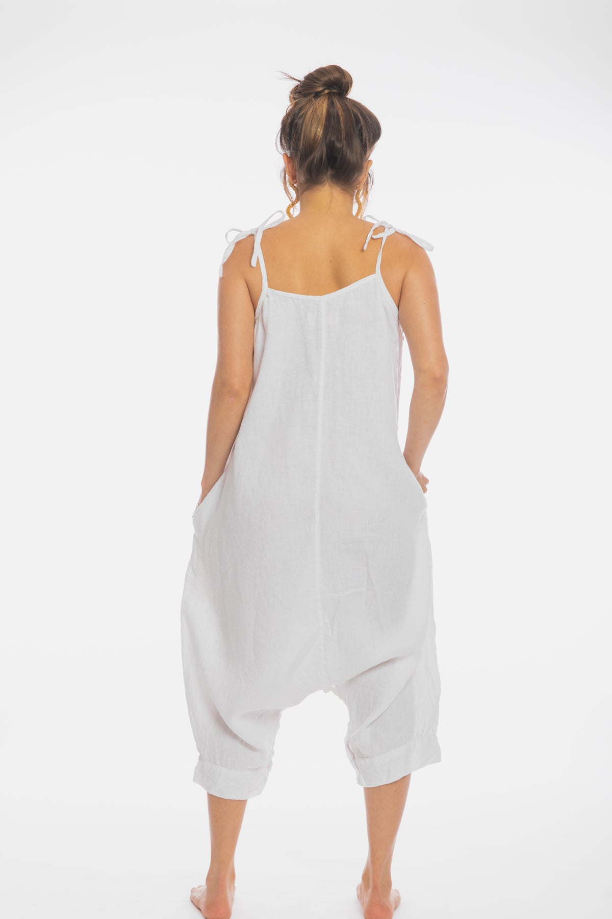 A person stands facing away from the camera, wearing a loose-fitting Japa Playsuit (100% Linen) WI by Myrah Penaloza with adjustable shoulder straps. The jumpsuit, crafted from linen fabric, has a casual and relaxed design, with the person’s hands in the pockets. Their hair is styled in a messy bun.