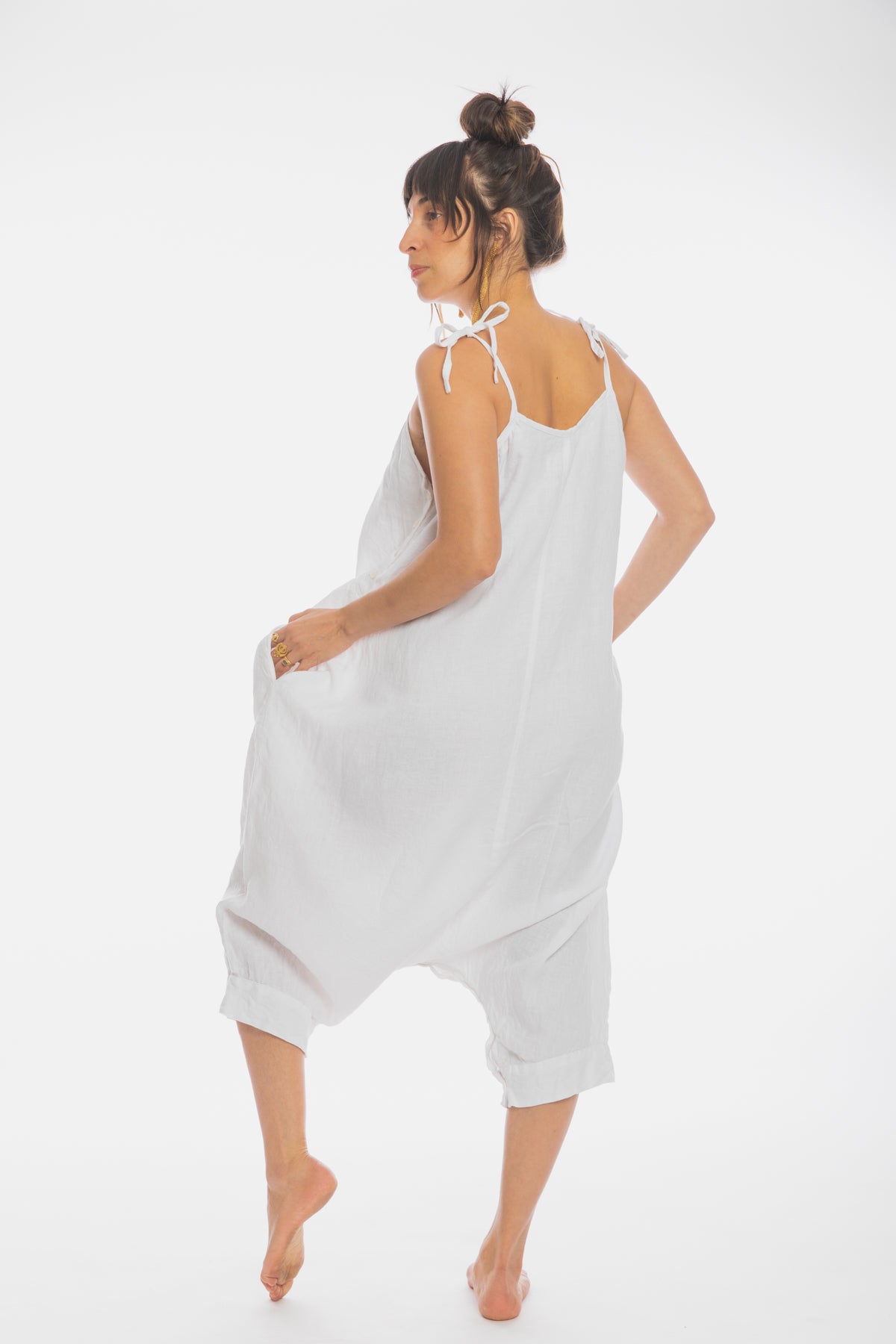 A person, barefoot against a white background, is wearing the Myrah Penaloza Japa Playsuit (100% Linen) WI with shoulder ties. They are looking over their shoulder with one hand in their pocket and have a high bun hairstyle. Their slightly bent leg conveys a casual, relaxed pose reminiscent of Kundalini Yoga serenity.